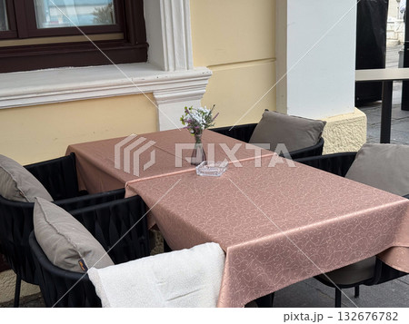 Outdoor cafe table with pink tablecloth and flower vase. Cozy urban terrace, relaxation, and street dining ambiance. 132676782
