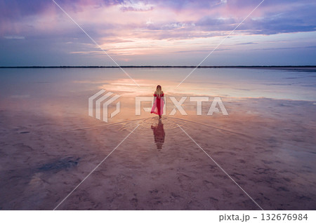Aerial of a young woman in red dress walking in the water of a unique pink salt lake. Sunset at lake Bursol with beautiful reflections on calm water surface. Stunning scenery 132676984