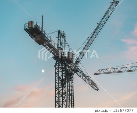 Crane silhouettes against pastel twilight sky with moon over construction site during evening Crane silhouettes against pastel twilight sky with moon over construction site during evening 132677077