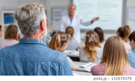 Rear view of adult student with gray hair listening to professor during classroom lecture in university setting Rear view of adult student with gray hair listening to professor during classroom lecture in university setting 132678379