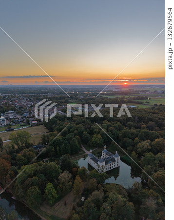 Aerial view at sunset white castle like building with moat and towers nestled in dense forest, winding path leads to entrance, warm sky glows over nearby town blending history, nature, and modern life 132679564