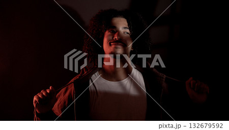 Young latin african-american man with curly hair and goatee is opening his brown hoodie in a dark room with red light Young latin african-american man with curly hair and goatee is opening his brown hoodie in a dark room with red light 132679592