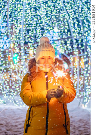 Young woman in knitted hat holds sparkler in her hands. Night festive street, shiny city decorations for Christmas. 132681914