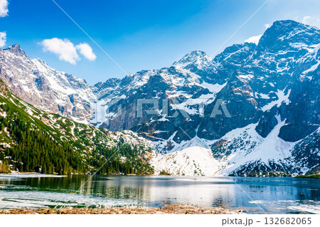 mountain lake in tatra national park. morskie oko surrounded with rugged snow capped peaks in early spring. beautiful view of reflection and ice on water. green environment background under blue sky mountain lake in tatra national park. morskie oko surrounded with rugged snow capped peaks in early spring. beautiful view of reflection and ice on water. green environment background under blue sky 132682065