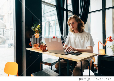 Young man working on laptop in a modern cafe setting with chess pieces displayed Young man working on laptop in a modern cafe setting with chess pieces displayed 132682657