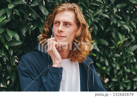 Man with wavy hair poses thoughtfully against a green leafy background in daylight 132682679