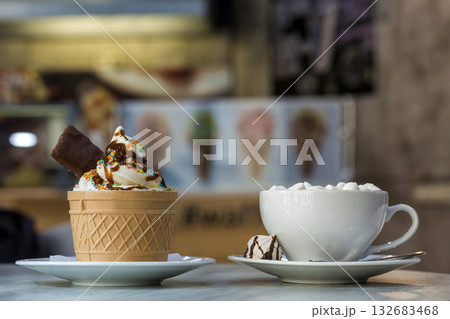 Ice cream dessert in wafer cup with chocolate cookies and and mug of coffee with marshmallows on porcelain plate on blurred colorful copy space background. Ice cream dessert in wafer cup with chocolate cookies and and mug of coffee with marshmallows on porcelain plate on blurred colorful copy space background. 132683468