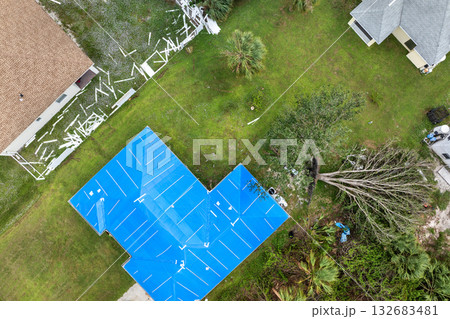 Hurricane affected house roof with blue tarp stretched over broken area to guard against rain until full asphalt shingle restoration can be carried out Hurricane affected house roof with blue tarp stretched over broken area to guard against rain until full asphalt shingle restoration can be carried out 132683481