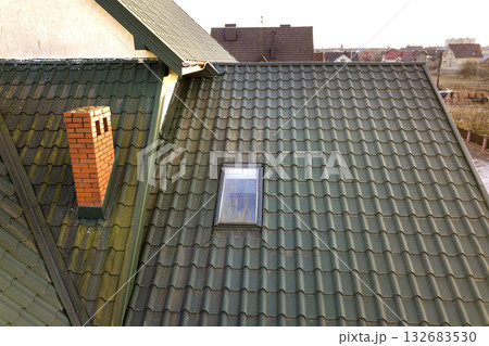 Green metal shingled house roof with attic plastic window and brick chimney. 132683530