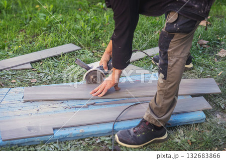 Worker Cutting Tile Outdoors. Construction worker cutting tile with electric grinder on grass field during outdoor renovation project. Concept of craftsmanship, renovation process, skilled labor 132683866