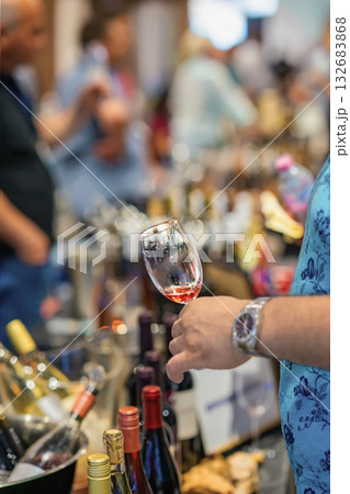 Man holds glass of wine, wine tasting event, surrounded by bottles and people, expo atmosphere. Concept of wine culture, social service gathering 132683868