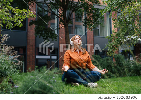 Beautiful mid size woman meditating in park, listening podcast. 132683906