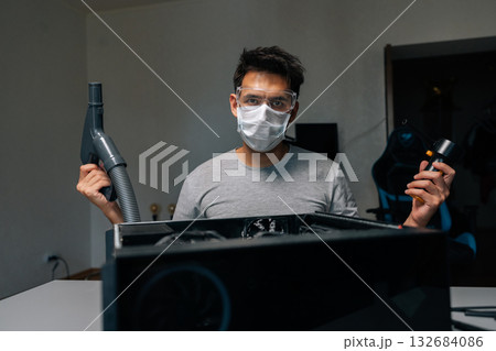 Portrait of young computer technician wearing protective mask and glasses, cleaning computer case using vacuum cleaner and blower, performing maintenance and ensuring optimal performance. 132684086