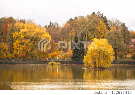 This tranquil pond in Kudowa Zdroj features vibrant autumn colors with shades of orange and yellow. Visitors stroll along the water's edge, enjoying the peaceful fall atmosphere. 132685410