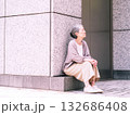 ビルの壁に腰掛けて遠くを見る白髪交じりのシニア女性 132686408