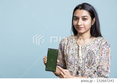 Happy indian woman presenting green screen on her smartphone, posing against the blue background. Joyous girl holding copy space device used for advertising brands in a commercial. Happy indian woman presenting green screen on her smartphone, posing against the blue background. Joyous girl holding copy space device used for advertising brands in a commercial. 132687241
