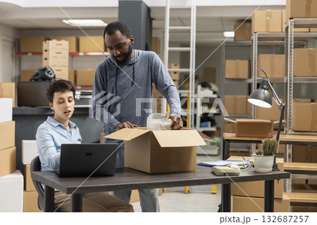 Employees team coordinating product packaging and local shipment in a supply room filled with boxes and goods, showing the daily workflow in a small scale logistics operation. Employees team coordinating product packaging and local shipment in a supply room filled with boxes and goods, showing the daily workflow in a small scale logistics operation. 132687257