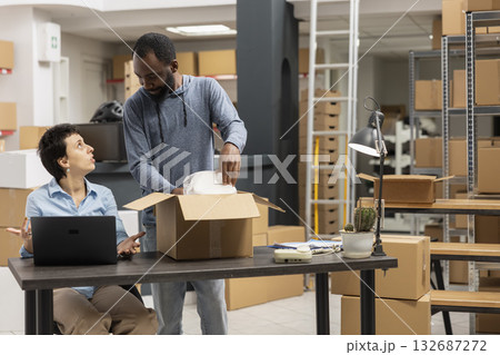 Young team of workers processing packages on a laptop in a busy shipping area, handling custom orders and shipment logistics with taped boxes. Small business local brand environment. Young team of workers processing packages on a laptop in a busy shipping area, handling custom orders and shipment logistics with taped boxes. Small business local brand environment. 132687272