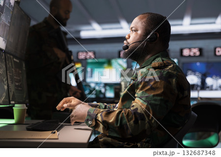 Military agent using headset microphone, offering information to commanders fighting on front lines. African american army specialist providing intel for troops in base of operations Military agent using headset microphone, offering information to commanders fighting on front lines. African american army specialist providing intel for troops in base of operations 132687348