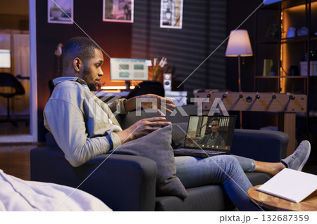 Focused black guy smiling and waving during a web conference in his living room, showcasing online discussion in a digital and wireless lifestyle. Conference speaker with attendees on call. Focused black guy smiling and waving during a web conference in his living room, showcasing online discussion in a digital and wireless lifestyle. Conference speaker with attendees on call. 132687359