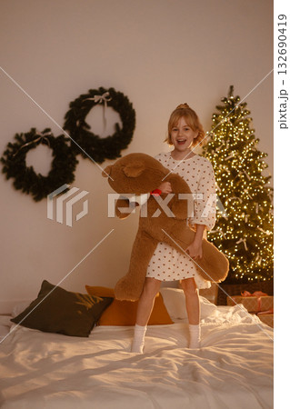 Girl hugging teddy bear with Santa hat front view sitting on bed near Christmas tree with lights 132690419