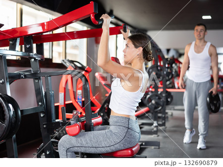 Girl doing strength training in gym, exercising upper body muscles on shoulder press machine Girl doing strength training in gym, exercising upper body muscles on shoulder press machine 132690879