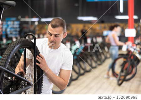 Young male customer choosing bike and checking tire on bicycle wheel in sport store 132690968