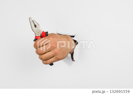 Hand holding pliers reaches through a torn wall in a playful scene Hand holding pliers reaches through a torn wall in a playful scene 132691559