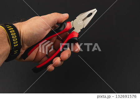 Hand holding red and black pliers against a dark background Hand holding red and black pliers against a dark background 132691576