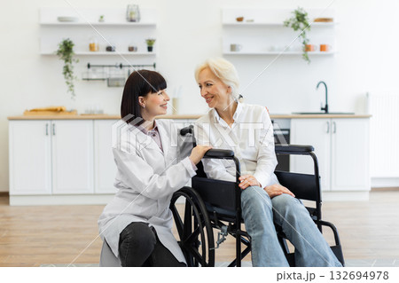 A smiling caregiver comforts an elderly woman in a wheelchair, fostering a sense of support and care in a bright, modern home setting. A smiling caregiver comforts an elderly woman in a wheelchair, fostering a sense of support and care in a bright, modern home setting. 132694978