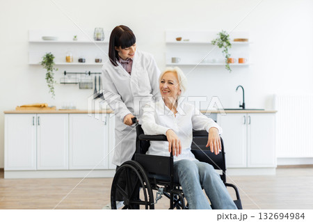 A smiling caregiver pushes an elderly woman in a wheelchair indoors, likely in a kitchen setting, offering support and care. 132694984