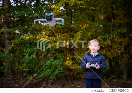 A joyful boy stands focused, controlling a drone amidst colorful autumn leaves. The sun shines, creating a cheerful atmosphere as nature surrounds him. 132698906