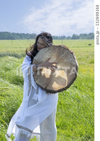 Female shaman in the white dress drumming outdoors 132699168