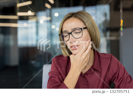 Woman wearing glasses touching her jaw with a hand while suffering from severe toothache pain, discomfort, and inflammation, disrupting her concentration at work in a modern office Woman wearing glasses touching her jaw with a hand while suffering from severe toothache pain, discomfort, and inflammation, disrupting her concentration at work in a modern office 132705751