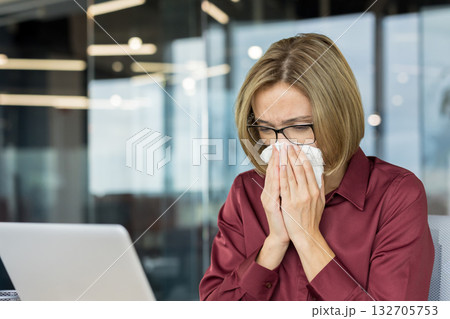 Young woman feeling unwell and blowing her nose with a tissue while working at her desk in a modern office, experiencing symptoms of cold, flu, or seasonal allergies Young woman feeling unwell and blowing her nose with a tissue while working at her desk in a modern office, experiencing symptoms of cold, flu, or seasonal allergies 132705753
