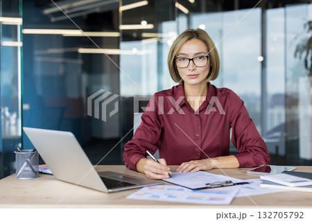Confident asian businesswoman in modern corporate office reviewing documents at her desk with laptop and glasses, poised and focused on financial planning and contract signing 132705792