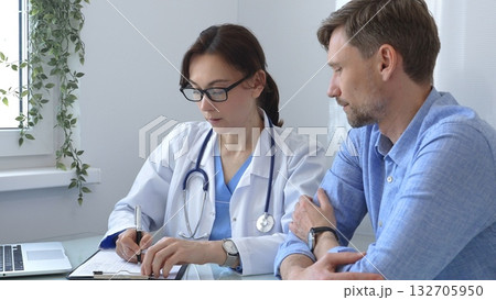 Female doctor consulting with patient in medical office using laptop and filling out paperwork to provide healthcare advice and support, discussing treatment options in a professional setting. Medical Female doctor consulting with patient in medical office using laptop and filling out paperwork to provide healthcare advice and support, discussing treatment options in a professional setting. Medical 132705950
