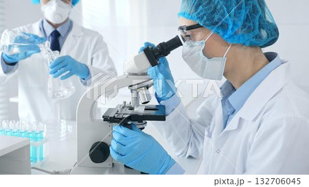 Masked female scientist examining specimen under microscope while laboratory assistant handles glassware in medical investigation at the background. Medicine, and science concept 132706045