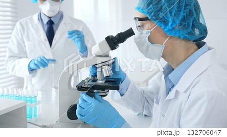 Woman scientist in blue protective gear is examining specimen under microscope while laboratory assistant handles glassware in medical investigation at the background. Medicine, and science concept 132706047