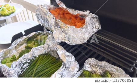 Grilled carrots, asparagus, and broccoli cook in foil packets on a gas grill with a white plate set nearby. The bright daylight suggests a relaxed summer setting. 132706851
