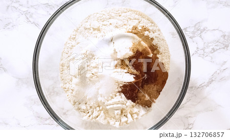 An overhead view of flour, sugar, and spices arranged together in a glass mixing bowl on a marble surface. The mixture highlights dry ingredient preparation for baking cookies. 132706857