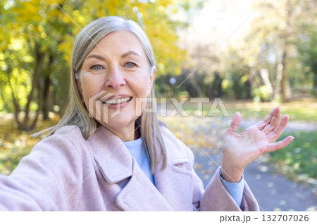 Senior woman smiling and waving hand during a video call or making a selfie video in an autumn park, embracing digital connectivity and communication with friends or family 132707026