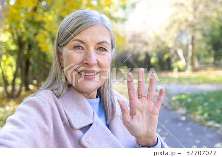 Senior woman smiling and waving her hand, communicating via video call or selfie in an outdoor autumn park, showing connection, technology, and friendly greeting Senior woman smiling and waving her hand, communicating via video call or selfie in an outdoor autumn park, showing connection, technology, and friendly greeting 132707027