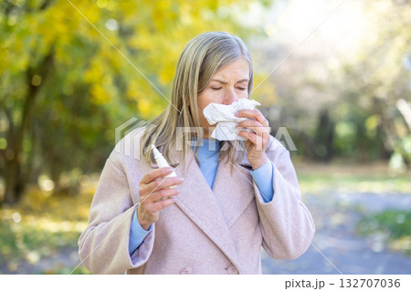 Senior woman with seasonal allergies in autumn park, blowing her nose into a tissue while holding a nasal spray, surrounded by fallen leaves and trees, seeking relief from symptoms 132707036