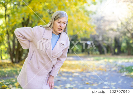 Senior woman standing bent over in an autumn park, suffering from acute lower back pain, clutching her side while showing a pained facial expression due to discomfort and health issues Senior woman standing bent over in an autumn park, suffering from acute lower back pain, clutching her side while showing a pained facial expression due to discomfort and health issues 132707040