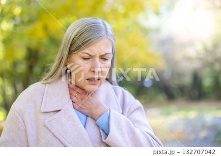Senior woman clutching her throat in pain, looking unwell in a chilly autumn park, showing symptoms of sore throat, cold or seasonal flu and discomfort Senior woman clutching her throat in pain, looking unwell in a chilly autumn park, showing symptoms of sore throat, cold or seasonal flu and discomfort 132707042