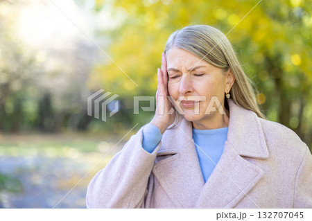 Mature woman squeezing her temples with eyes closed, expressing discomfort and pain from a headache or migraine while standing in an autumn park with blurred yellow foliage Mature woman squeezing her temples with eyes closed, expressing discomfort and pain from a headache or migraine while standing in an autumn park with blurred yellow foliage 132707045