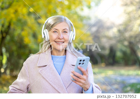Senior woman smiling and listening to music through headphones, holding a smartphone while relaxing outdoors in an autumn park, embracing technology and leisure Senior woman smiling and listening to music through headphones, holding a smartphone while relaxing outdoors in an autumn park, embracing technology and leisure 132707052