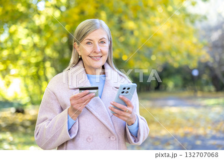 Happy senior woman standing in an autumn park, looking at the camera and smiling while holding a credit card and a smartphone for online shopping and e-commerce transactions Happy senior woman standing in an autumn park, looking at the camera and smiling while holding a credit card and a smartphone for online shopping and e-commerce transactions 132707068