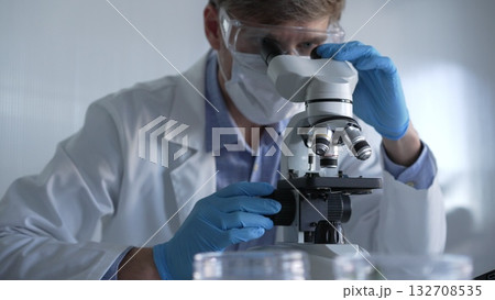 Male scientist researcher wearing blue gloves, protective glasses and medical mask, is using microscope in modern laboratory, portrait view. Low key lighting photo. Science and Medicine Male scientist researcher wearing blue gloves, protective glasses and medical mask, is using microscope in modern laboratory, portrait view. Low key lighting photo. Science and Medicine 132708535